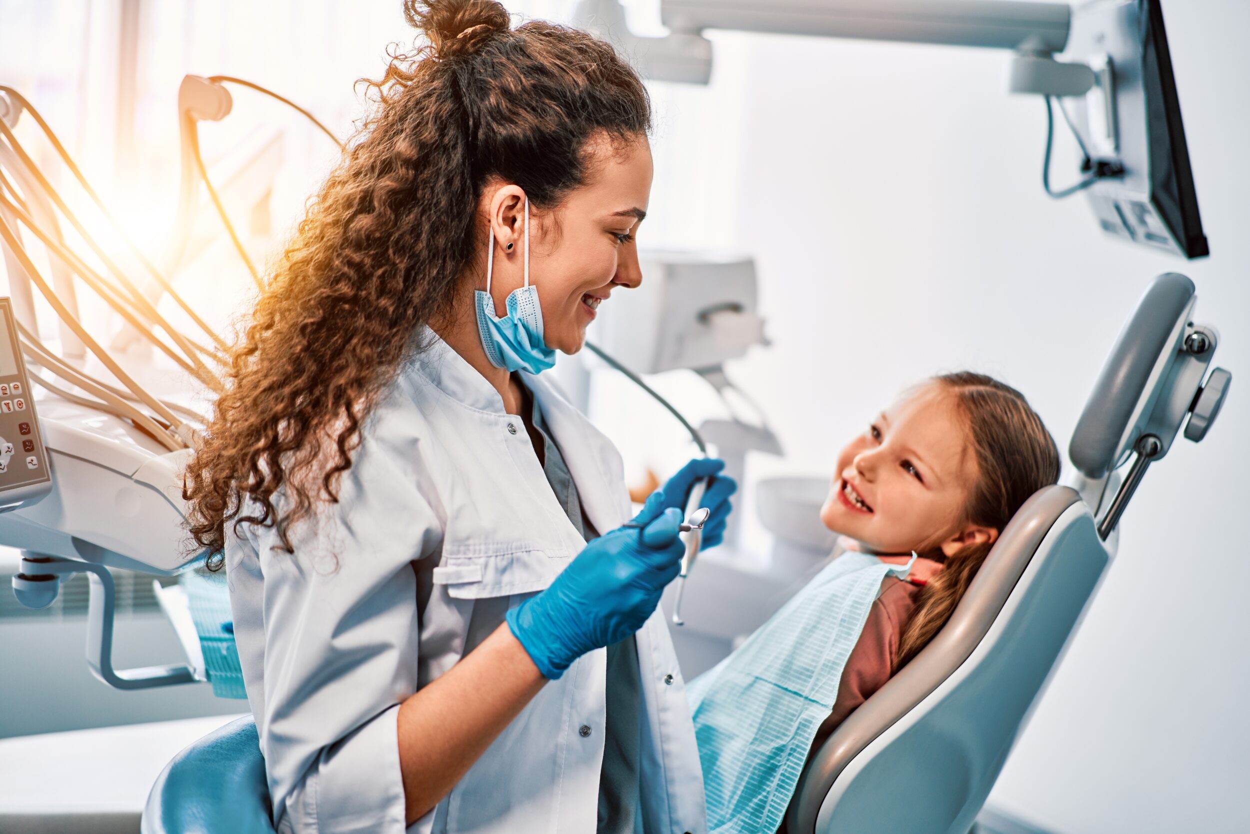 Female dentist examining smiling child’s teeth in dental chair with sunlight, discussing white composite fillings