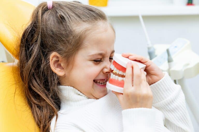 Young girl sitting in dental office holding a jaw model, making faces while learning about teeth, representing special needs pediatric dentistry and oral care education
