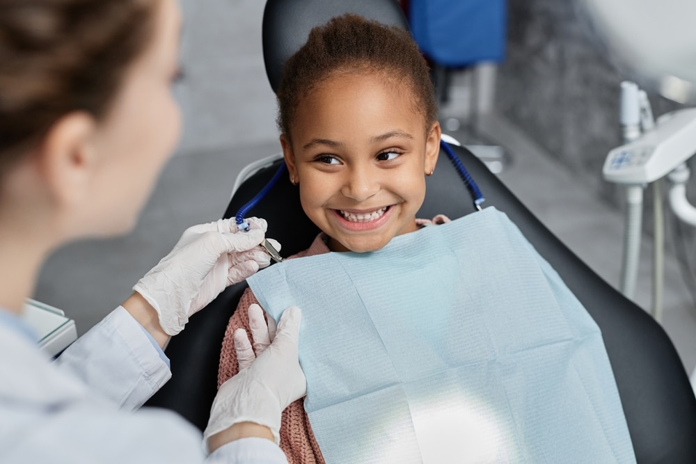 Smiling child sitting in dental chair as nurse prepares for teeth checkup and oral health care.