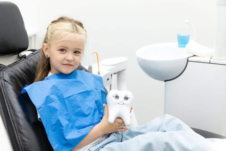 Happy child sitting in dental chair holding artificial tooth model at pediatric dental clinic, concept of fun and friendly children’s dentistry.