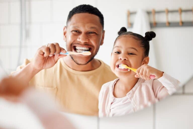 Father and young daughter brush their teeth together in front of the bathroom mirror.