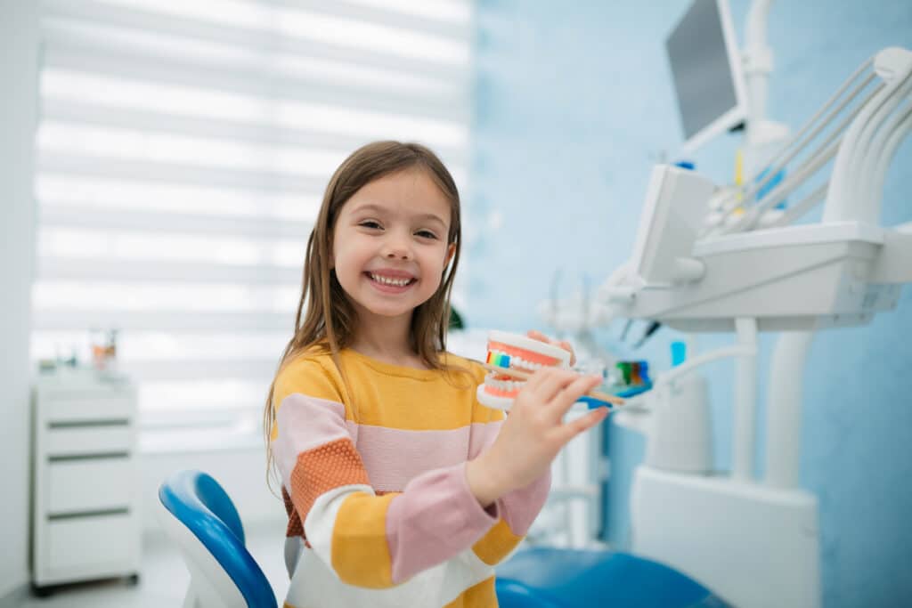 Smiling young girl holding a dental model and toothbrush in a pediatric dentist’s office.