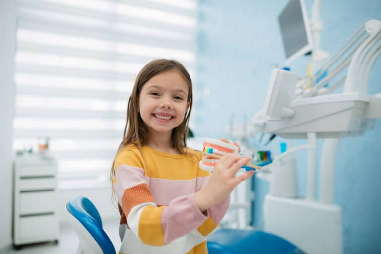 Smiling young girl holding a dental model and toothbrush in a pediatric dentist’s office.
